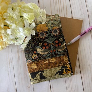 May include: A patterned journal cover with a brown card and a pink pen. The journal cover has a black background with birds, flowers, and berries in brown, green, and yellow. A bouquet of yellow and white flowers is in the upper left corner.