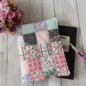 May include: A quilted fabric book cover featuring a patchwork design of floral and geometric patterns in shades of pink, teal, and white. The cover is placed over a black notebook with white polka dots, accompanied by a clear pen with a pink accent.