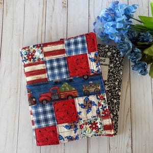 Peut inclure: Une couverture de carnet en tissu patchwork rouge, blanc et bleu avec un motif de camion rouge et de pastèque. La couverture est pliée en deux et repose sur une surface en bois blanc. Un carnet de composition noir et blanc est visible sous la couverture.