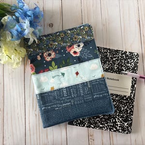 May include: A fabric book cover featuring horizontal panels of floral, kite, and typography prints in shades of blue, white, and pink. The cover is placed over a black and white speckled composition notebook with a pink and white ballpoint pen.