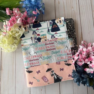 May include: A colorful fabric book cover with a patchwork design featuring a repeated pattern of the words "Super Expialafragi Califragi docious" and a silhouette of a woman with an umbrella. The cover is made of multiple fabrics with different patterns and colors, including pink, blue, green, and white.