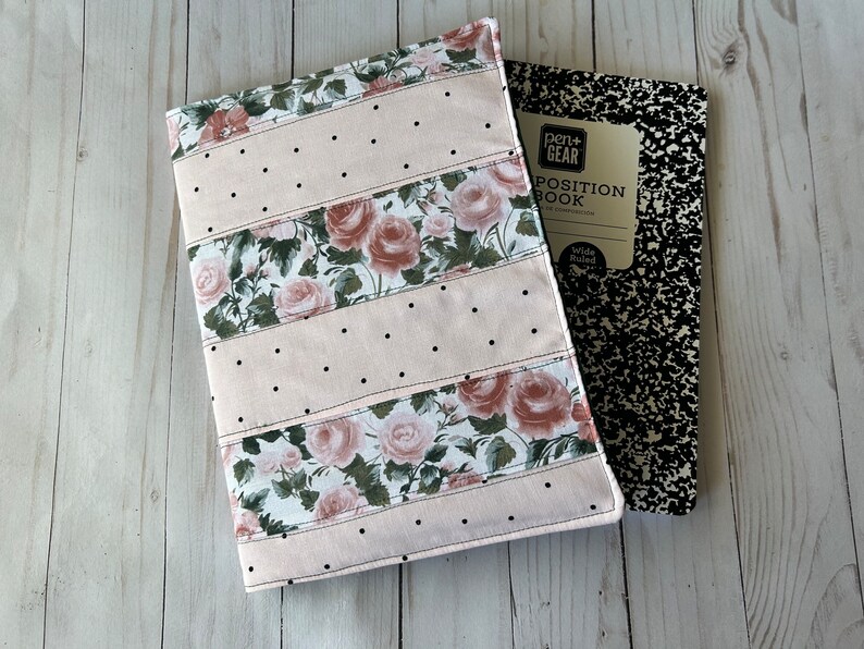 May include: A floral notebook cover with a pink background and black polka dots, paired with a composition notebook. The cover features rose patterns and is stitched with black thread. The composition notebook has a black and white speckled design.