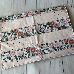 May include: A rectangular, floral-patterned placemat. The design features alternating stripes of blush pink fabric with black polka dots and fabric with a rose pattern. The edges are finished with a stitched border.