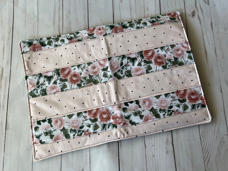 May include: A rectangular, floral-patterned placemat. The design features alternating stripes of blush pink fabric with black polka dots and fabric with a rose pattern. The edges are finished with a stitched border.