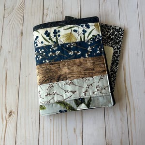 May include: A quilted notebook cover with a patchwork design. The cover features various fabric patterns, including floral, wood grain, and botanical prints in shades of blue, white, and brown. A black and white composition notebook is visible inside.
