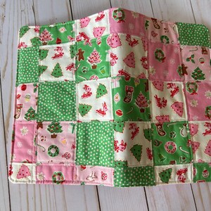 May include: A patchwork Christmas-themed item featuring squares of green, pink, and white fabric. The squares are decorated with Christmas trees, gingerbread men, wreaths, and polka dots. The item is likely a placemat or small quilt.