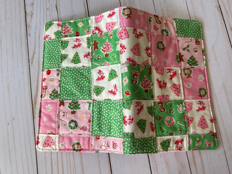 May include: A patchwork Christmas-themed item featuring squares of green, pink, and white fabric. The squares are decorated with Christmas trees, gingerbread men, wreaths, and polka dots. The item is likely a placemat or small quilt.