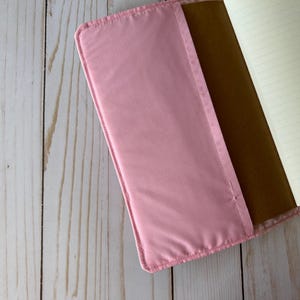 May include: A pink notebook cover with a soft, padded texture. The cover is open, revealing a brown interior and lined paper pages. The notebook is set against a light wood grain background.