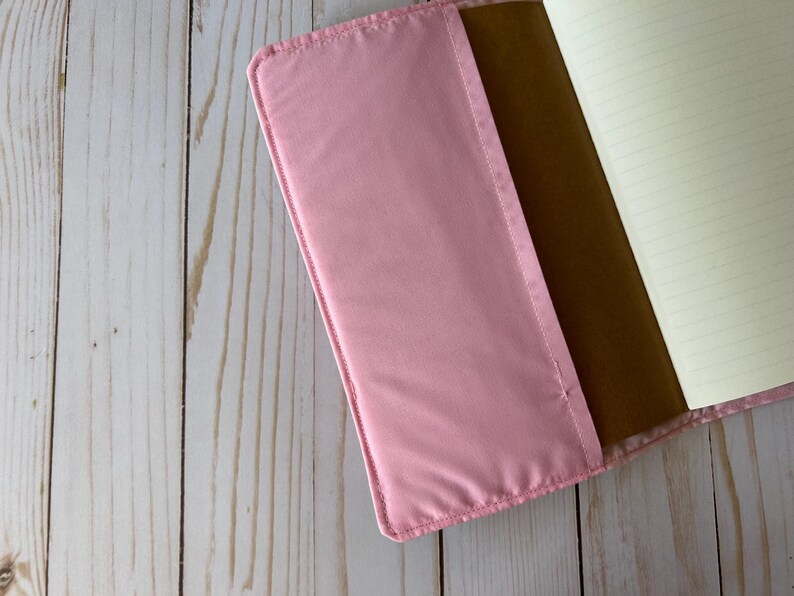 May include: A pink notebook cover with a soft, padded texture. The cover is open, revealing a brown interior and lined paper pages. The notebook is set against a light wood grain background.