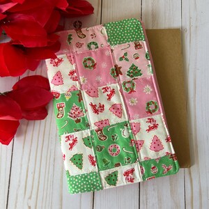 May include: A patchwork Christmas-themed tablet cover. The cover features a mix of pink, green, and white squares with festive patterns like Christmas trees, wreaths, and stockings. The cover is placed on a light wooden surface, with red flowers in the background.