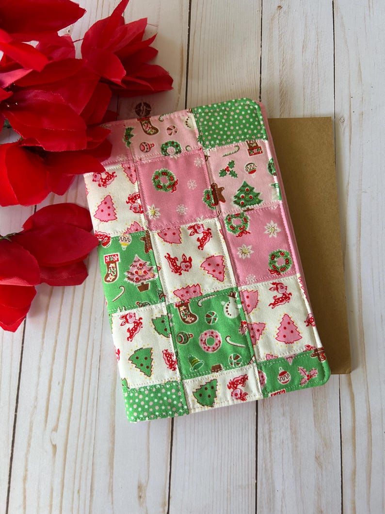 May include: A patchwork Christmas-themed tablet cover. The cover features a mix of pink, green, and white squares with festive patterns like Christmas trees, wreaths, and stockings. The cover is placed on a light wooden surface, with red flowers in the background.