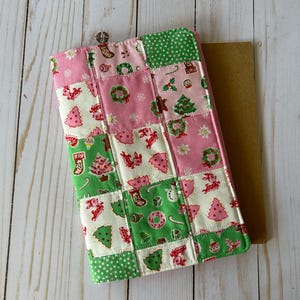 May include: A patchwork Christmas-themed tablet cover featuring squares of pink, green, and white fabric. The fabric is decorated with festive images like Christmas trees, wreaths, and stockings. The cover is rectangular and has a brown backing.