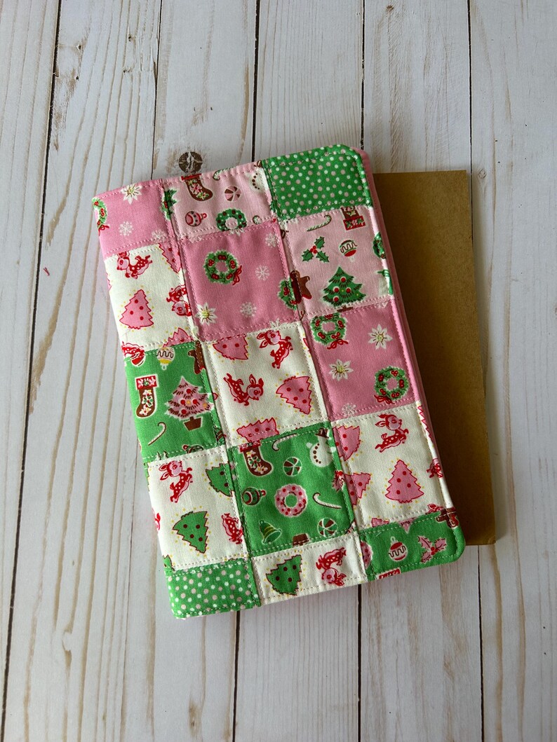 May include: A patchwork Christmas-themed tablet cover featuring squares of pink, green, and white fabric. The fabric is decorated with festive images like Christmas trees, wreaths, and stockings. The cover is rectangular and has a brown backing.