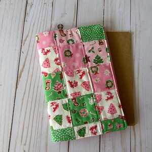 May include: A patchwork Christmas-themed tablet case. The case features a variety of festive patterns, including Christmas trees, wreaths, and stockings, in pink, green, and white. The case is rectangular and appears to be made of fabric.