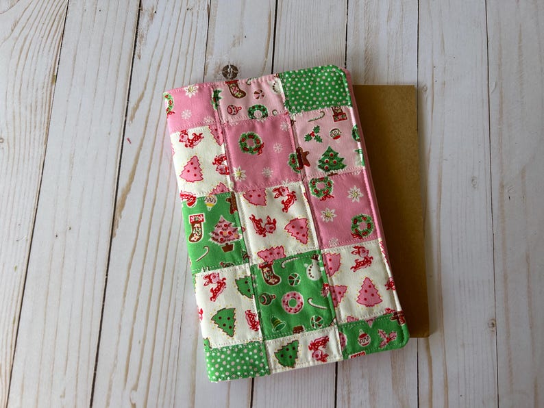 May include: A patchwork Christmas-themed tablet case. The case features a variety of festive patterns, including Christmas trees, wreaths, and stockings, in pink, green, and white. The case is rectangular and appears to be made of fabric.