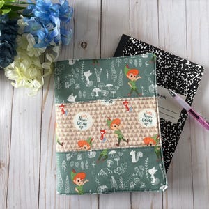 Peut inclure: Une couverture de carnet en tissu avec un fond vert orné d'illustrations blanches de Peter Pan, d'arbres et d'animaux des bois. Une bande horizontale centrale présente un motif de triangles beige et le texte Let's Never Grow Up.