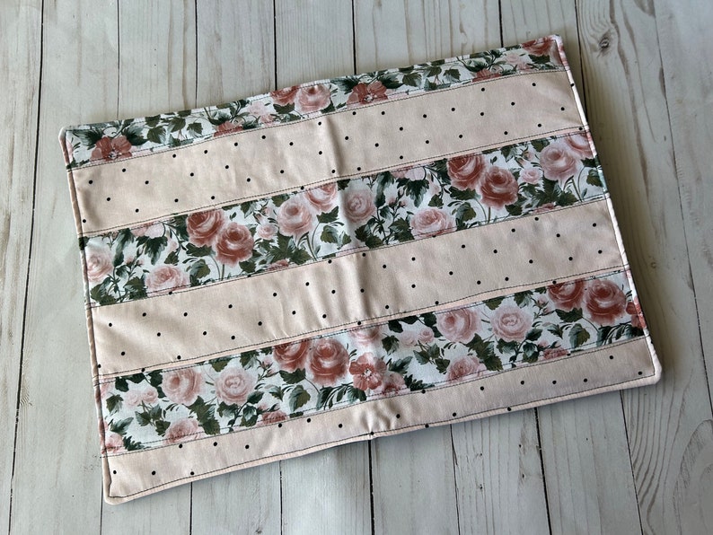 May include: A rectangular, floral-patterned placemat. The design features alternating stripes of blush pink fabric with black polka dots and fabric with a rose pattern. The edges are finished with a stitched border.