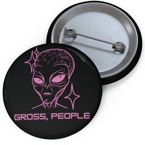 May include: Two black round buttons. One button features a pink alien illustration with the text "GROSS, PEOPLE". The other button is silver. The alien has large eyes and is surrounded by star shapes.