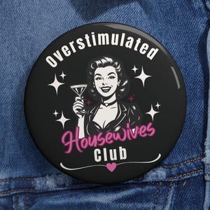 May include: A black button with the words "overstimulated" and "Housewives Club" in white and pink, respectively. The button features a vintage illustration of a woman holding a cocktail, with star accents.