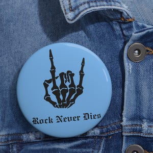 May include: A circular pinback button with a light blue background featuring a black graphic of a skeleton hand making a rock and roll hand gesture. The text "Rock Never Dies" is printed in a black gothic-style font at the bottom of the button.