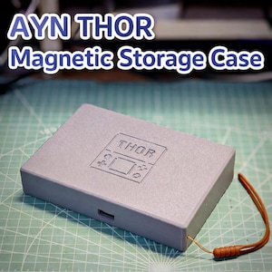 Puede incluir: Una caja de almacenamiento magnética rectangular gris con el texto "AYN THOR Magnetic Storage Case" en blanco y azul. La caja tiene un pequeño diseño en relieve y una correa de muñeca marrón. La caja está sobre una superficie verde.