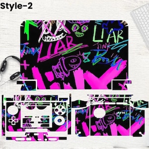 May include: A black gaming console skin with a graffiti-style design. The skin features neon pink, green, and white scribbled text and graphics, including the word "LIAR" and a cartoon character. The design covers the console and controller.