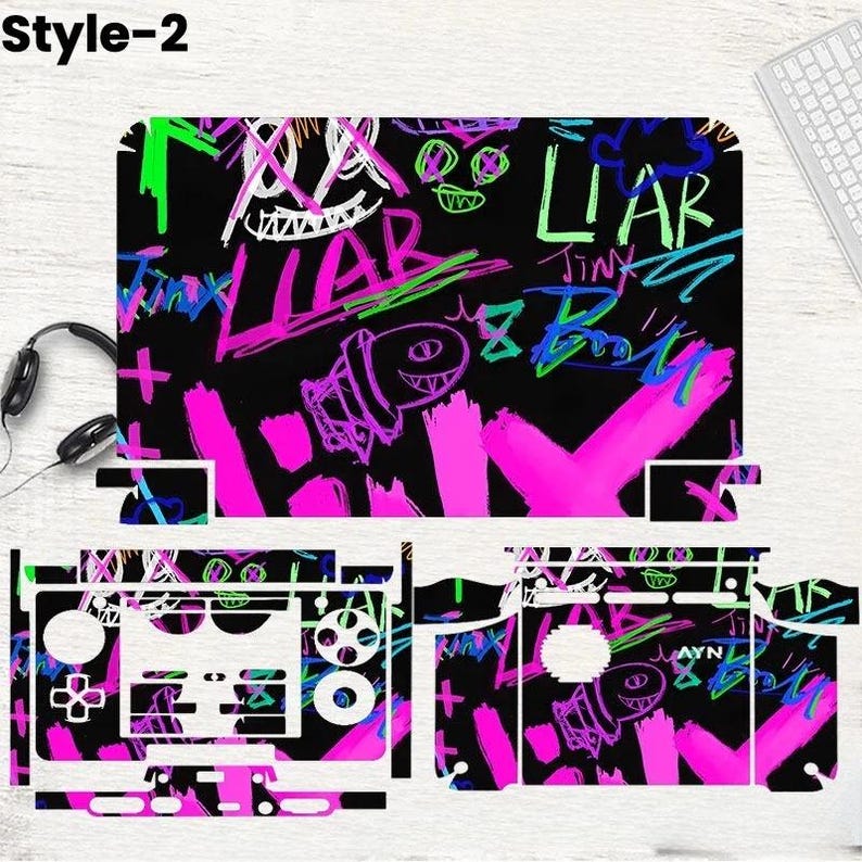 May include: A black gaming console skin with a graffiti-style design. The skin features neon pink, green, and white scribbled text and graphics, including the word "LIAR" and a cartoon character. The design covers the console and controller.