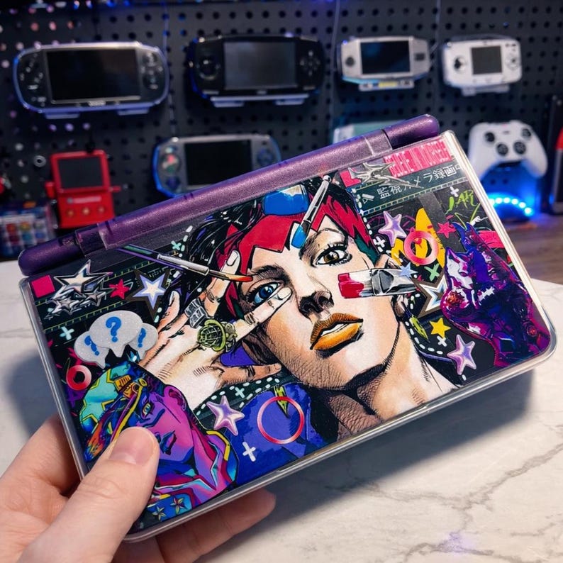 Puede incluir: Consola de juegos port&aacute;til con un dise&ntilde;o gr&aacute;fico vibrante y colorido. El dise&ntilde;o presenta una ilustraci&oacute;n de estilo manga de una persona con cabello rojo y ojos azules. La consola tiene una bisagra morada y se sostiene en una mano.