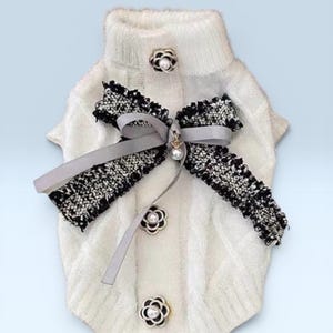 May include: A white dog sweater with a high collar, adorned with a black and white tweed bow and decorative flower-shaped buttons with pearl centers. The sweater has a cable knit design.