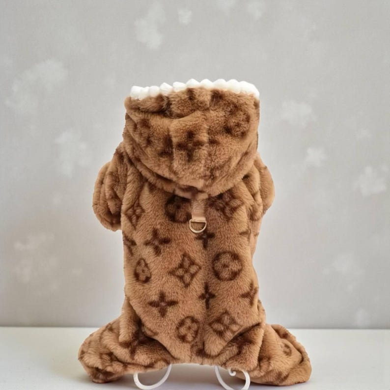 May include: Brown dog jumpsuit with a hood and a repeating logo pattern. The jumpsuit has a white trim around the hood and a small gold-colored ring on the back. The material appears to be soft and plush.