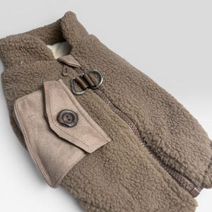 May include: A brown fleece jacket with a high collar and a full-length zipper. The jacket features a small pocket with a button closure and a silver-colored clasp. The material has a textured, fuzzy appearance.
