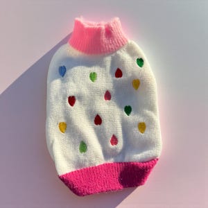 May include: A white pet sweater with a pink collar and hem, adorned with colorful embroidered hearts. The sweater has a turtleneck design and is made of a soft, textured material. The hearts are in various colors, including red, green, yellow, and blue.