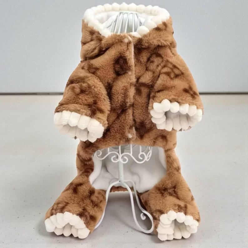May include: A brown and white pet jumpsuit with a hood and paw-like details. The jumpsuit has a plush, textured appearance with a button closure. The interior lining and paw tips are white.