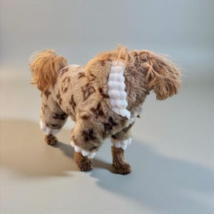 May include: A small dog wearing a brown and tan patterned outfit with white trim. The dog's fur is styled to match the outfit, and it stands on a light gray surface. The outfit has a repeating pattern.