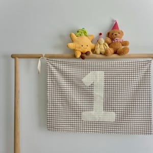 May include: A wooden frame holds a banner with a large number 1, made of light fabric with a brown and white gingham pattern. Above, plush toys including a star, a bunny, and a teddy bear with a party hat are displayed.