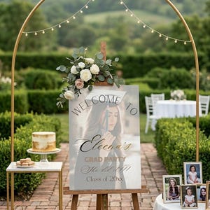 May include: A graduation party setup with a gold arch adorned with string lights. A sign on an easel reads "Welcome to Eleena's Grad Party, She did it!! Class of 20xx." A small cake and framed photos are also displayed.