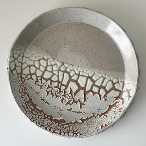 May include: A round ceramic plate featuring a speckled light gray finish on the upper half and a textured, cracked white glaze over a dark brown base on the lower half, creating a distinct contrast in surface patterns and color.