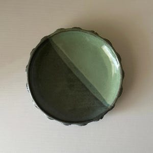 May include: A handcrafted ceramic plate with a distinctive design. The plate is split diagonally into two sections, one in a darker green and the other in a lighter green. The rim has a textured, uneven edge, enhancing its rustic appeal.