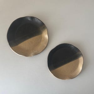 May include: Two circular ceramic dishes with an irregular, organic edge. Each dish features a two-tone design, with one half finished in a matte black glaze and the other half in a metallic gold finish, creating a distinct contrast between the two.
