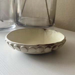 Op de afbeelding: Een kleine, gebroken witte keramische kom met een gestructureerd, cirkelvormig patroon rond de rand. De binnenkant heeft een lichtgele tint en de rand heeft een verweerde, bruine afwerking. De kom staat op een wit oppervlak.