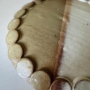 Op de afbeelding: Een handgemaakte keramische plaat met een uniek ontwerp. De plaat heeft een tweekleurig kleurenschema, met lichtbeige en bruine tinten. De rand is versierd met kleine, cirkelvormige accenten. De plaat heeft een textuur en een rustieke uitstraling.