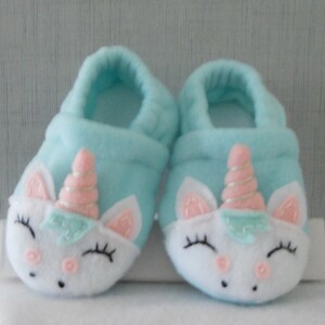 May include: A pair of light blue baby booties with a white unicorn design. The unicorn has pink ears, a pink horn with a gold swirl, and a light blue bow.