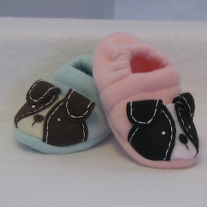 May include: A pair of baby booties with dog faces. One bootie is light blue with brown dog ears and a white nose. The other bootie is pink with black dog ears and a white nose.
