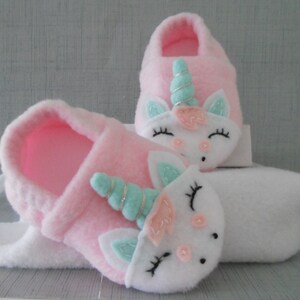 May include: A pair of pink fleece baby slippers with a white unicorn applique. The unicorn has a pink nose, blue eyes, and a light blue and silver horn.