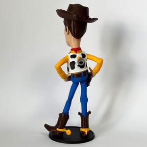 May include: A plastic figurine of a cowboy character wearing a brown hat, a white vest with black cow-print spots, a red neckerchief, yellow long sleeves, blue pants, and brown boots with yellow spurs. A pull-string ring is visible on the back.