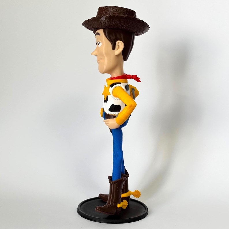 May include: A plastic figurine of a cowboy character wearing a brown hat, a yellow long-sleeved shirt, a white vest with black cow-print spots, a red neckerchief, blue pants, and brown boots with yellow spurs, standing on a round black base.
