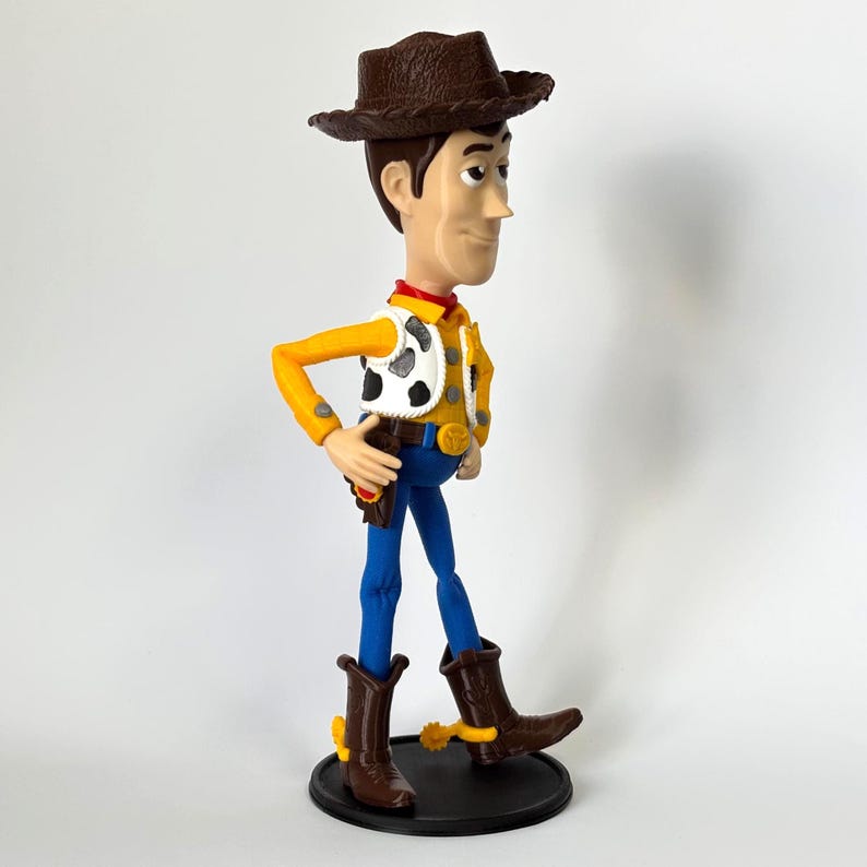May include: A standing cowboy figurine wearing a brown textured hat, a yellow long-sleeved shirt, a white vest with black cow-print spots, a red neckerchief, blue trousers, and brown boots with yellow spurs, all mounted on a circular black base.