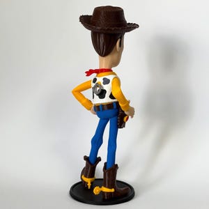 May include: A standing cowboy figurine featuring a brown cowboy hat, a white vest with black cow-print spots, a red neckerchief, blue pants, and brown boots with yellow spurs. A grey pull-string ring is visible on the back of the vest.