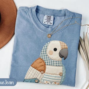 Puede incluir: Una camiseta color azul mezclilla con una aplicación grande de loro acolchado hecha de varios parches de tela con patrones de cuadros, rayas y flores. El ave tiene un pico gris oscuro, un ojo negro redondo y pequeños botones de madera.