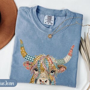 May include: A blue jean coloured t-shirt featuring a graphic of a highland cow with a patchwork pattern of floral and plaid designs in shades of yellow, pink, green, and white. The shirt has a crew neckline and a Comfort Colors brand label.